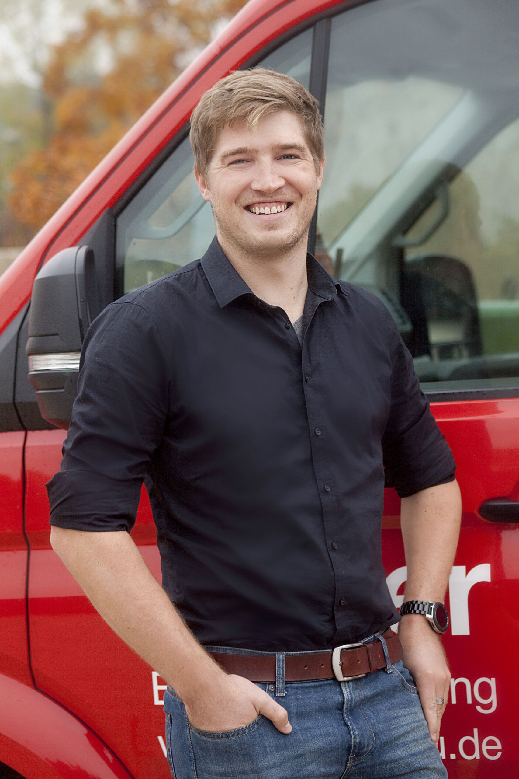 Manuel Schweizer: Vermessung. Mann vor rotem Fahrzeug, lässig.